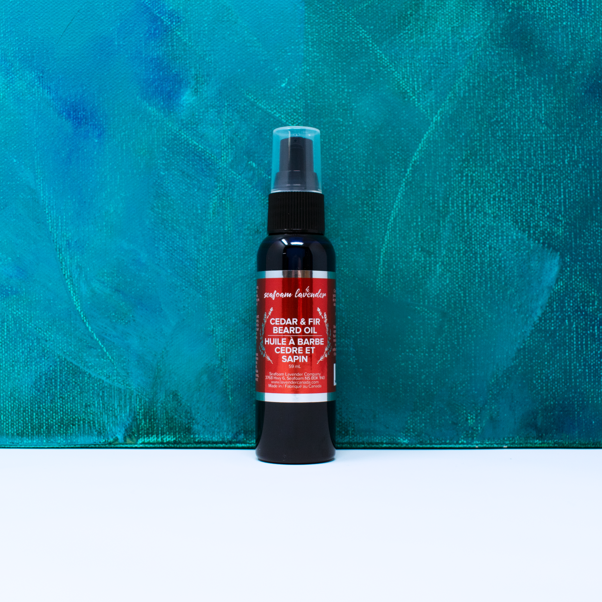 A crimson metallic label for Cedar & Fir Beard Oil on a dark blue bottle with a black pump top in front of a blue painted background. The label has plain sans serif text surrounded by a wreath of lavender sprigs.